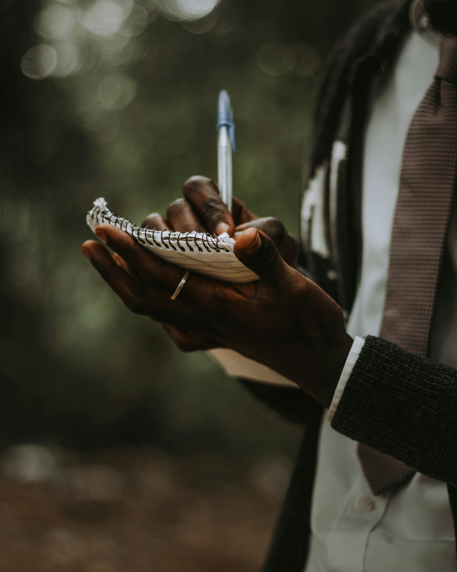 A person writing in a notebook outdoors, wearing a suit and tie, in soft natural light.